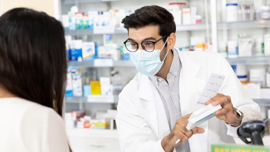 A pharmacist, wearing glasses, a blue surgical mask, and a white lab coat, explains a prescription to a patient with long dark hair.