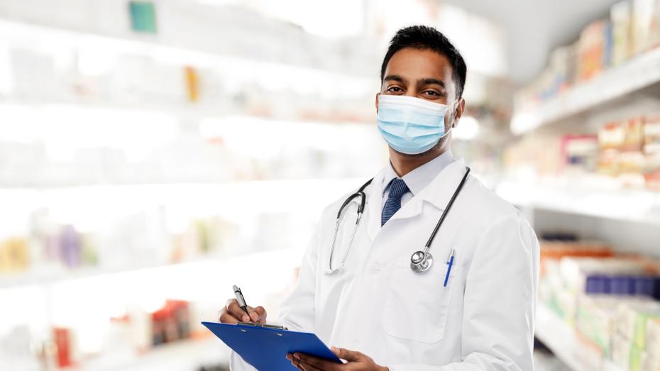 A pharmacist wearing a surgical mask and white coat stands holding a clipboard in a bright pharmacy stock room.